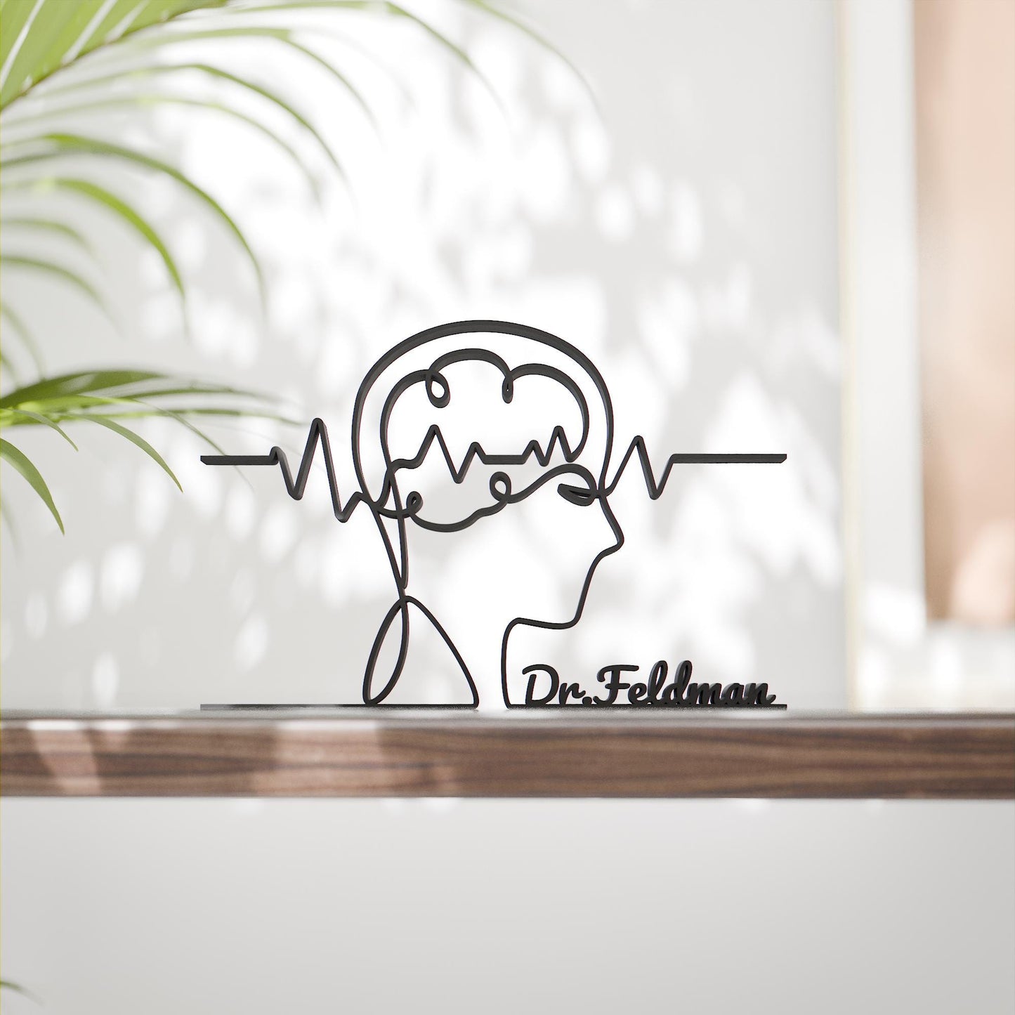 May include: Black metal sculpture depicting a human profile with a brain and a line representing brain activity. The sculpture includes the name "Dr. Feldman" at the base. The artwork is displayed on a wooden surface.