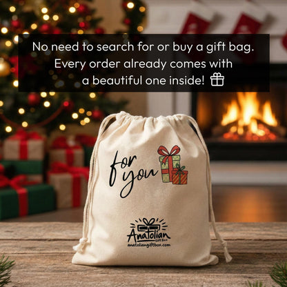 A beige drawstring bag with a gift bag design, placed on a wooden surface in front of a fireplace with Christmas lights and presents.